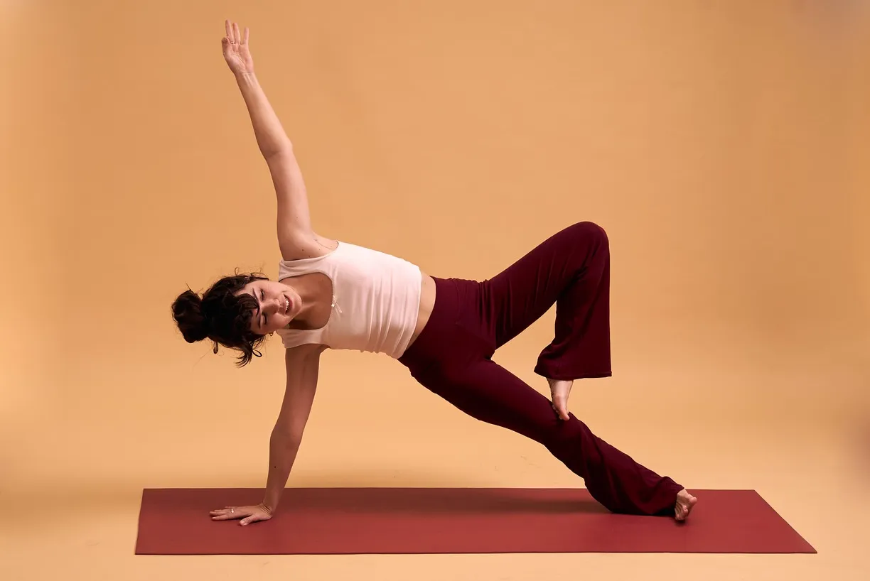 Mujer realizando una postura de yoga en una esterilla roja, con un fondo amarillo, levantando un brazo y manteniendo una pierna elevada, vestida con una camiseta blanca y pantalones rojos.